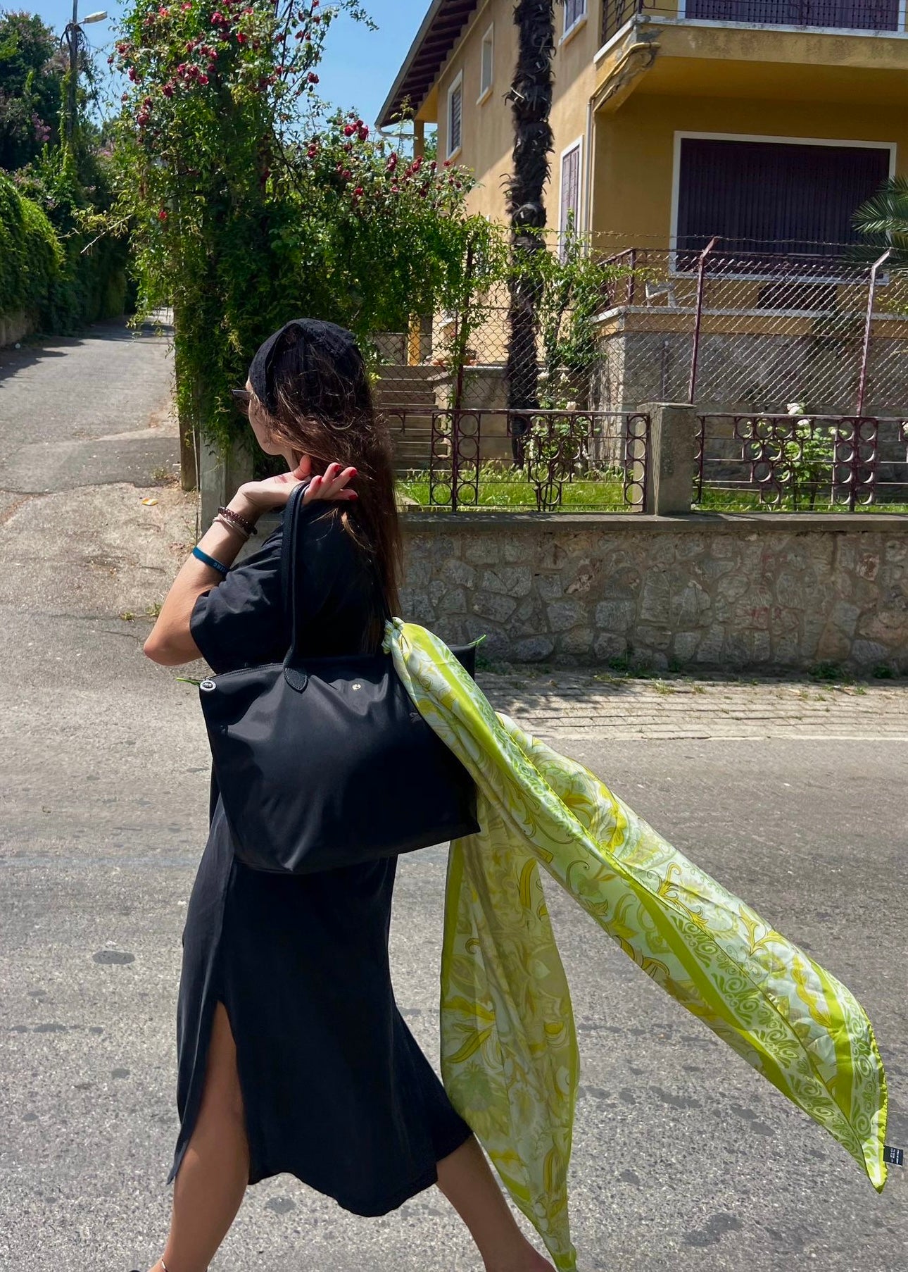 Woman carrying the Dorabella green and beige silk scarf hanging from her black bag while walking through a small town — effortless everyday styling of a luxury Italian scarf.