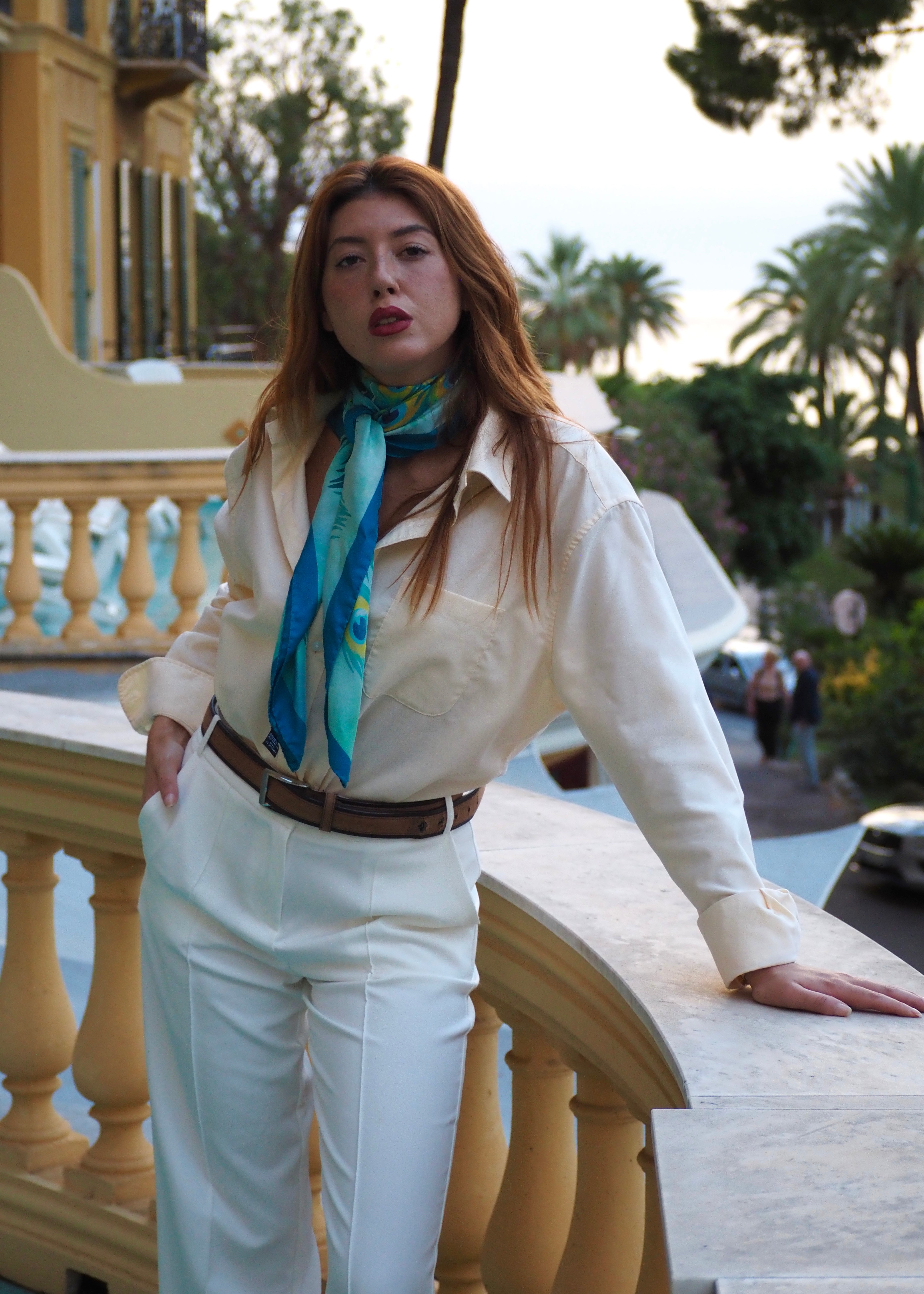 Woman wearing the Preziosa blue silk scarf with peacock design wrapped around her neck in Monte Carlo, Monaco — luxury Italian silk twill scarf for women.
